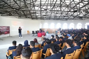 Speaker presenting on stage to a large audience in an auditorium with arched windows and exposed ceiling beams