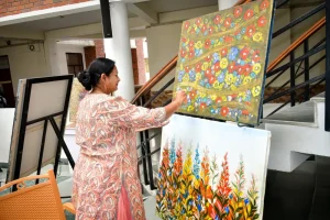 Woman painting a vibrant floral canvas in a studio, with another floral piece nearby on an easel outside a brick-walled building.