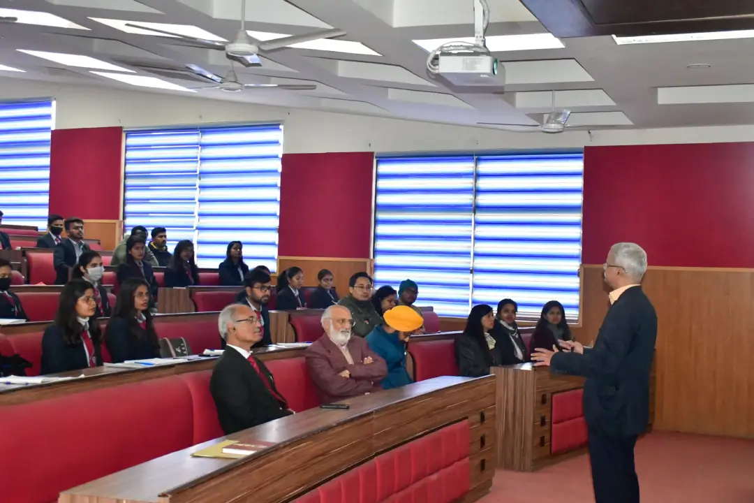 Speaker in a dark suit addresses a lecture hall filled with students seated in red seats.