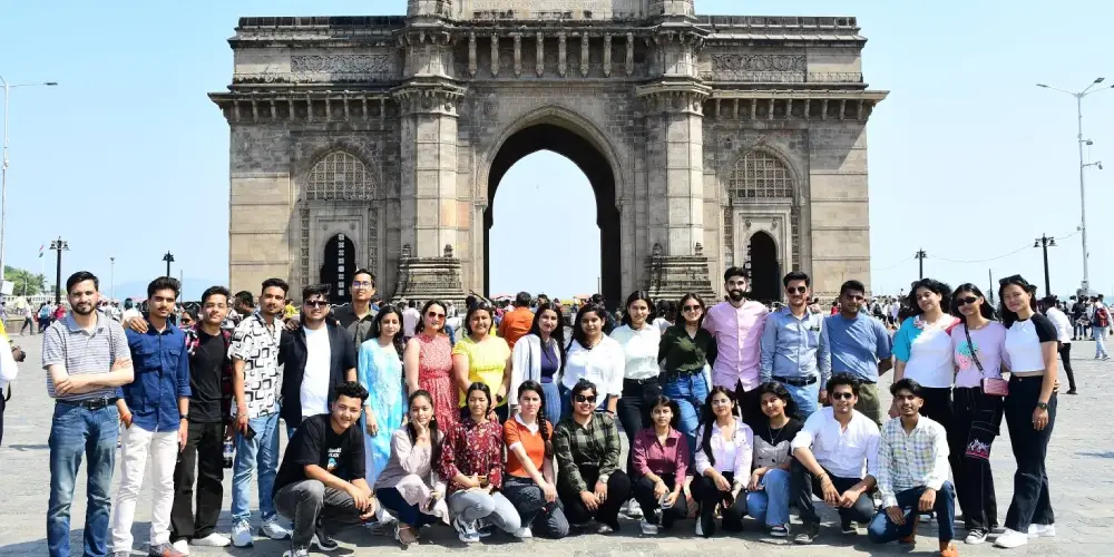 Group of young adults posing for a photo in front of the Gateway of India on a sunny day.