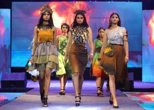 Group of models walking a runway in earth-toned dresses with bold headpieces on a colorful stage backdrop.