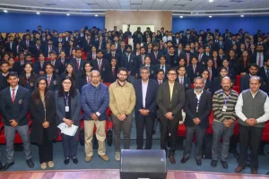 Group photo of students in uniforms with teachers and staff posing on stage in a blue-walled auditorium.