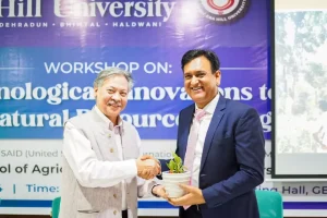 Two men in formal attire shake hands while one hands a small potted plant to the other at a workshop backdrop.
