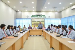 Group of students in uniforms seated around a U-shaped conference table, a man standing and addressing them at the center with a banner behind.