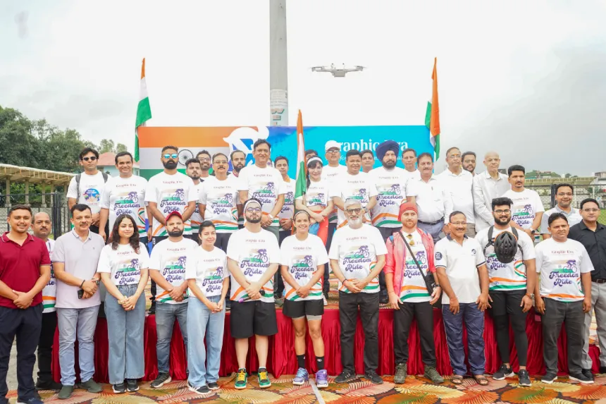 Group of people in white T-shirts posing on an outdoor stage with Indian flags and a drone overhead.