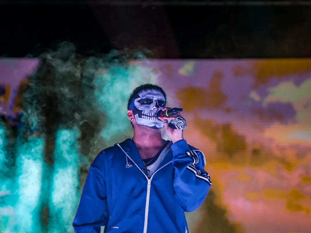 Performer wearing a blue jacket with skull makeup singing into a microphone on a foggy, colorfully lit stage.