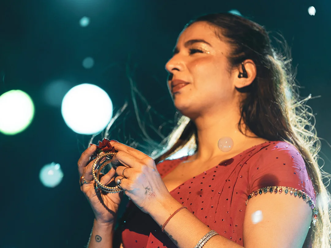 Female singer on stage wearing a pink dress, holding a tambourine with red ribbons, mic earpiece visible.