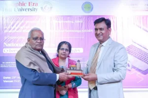Older man in a blue suit and scarf hands a trophy to a smiling man in a light gray suit on stage, with a woman in a red jacket between them and a purple banner in the background.