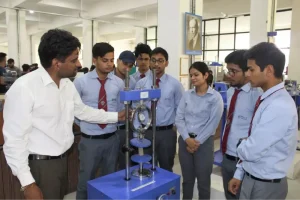 Instructor demonstrates a vertical mechanical testing device to a group of students in a lab classroom.