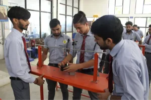 Group of students in shirts and ties work together on a red lab testing rig with a dial gauge in a classroom lab setting.