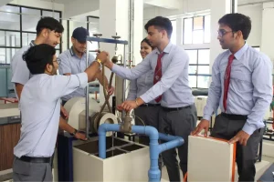 Engineering students in uniform collaborate around a lab bench, adjusting a pulley-and-rope setup with blue piping nearby.