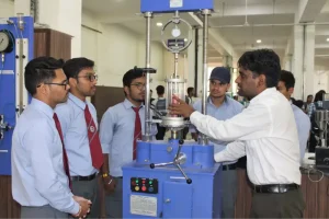 Instructor demonstrates a hydraulic press to a group of students in a lab, with blue testing equipment and gauges visible.