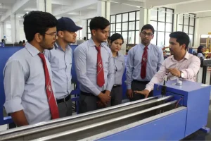 Five students in light blue shirts and red ties watch a man demonstrate a blue industrial machine in a workshop.