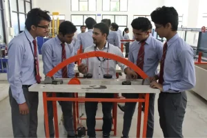 Group of students in school uniforms gathered around a lab bench with an orange circular frame, examining a mechanical demonstration in a workshop.