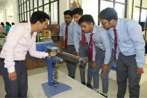 Instructor demonstrates a metalworking machine to five students in a workshop classroom, all in uniform shirts and ties.
