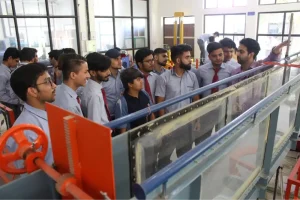 Group of students in blue shirts and red ties on a factory tour, listening to a guide beside an industrial machine with a red wheel in a workshop environment.