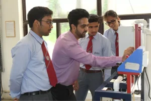 Instructor in a pink shirt demonstrates a machine to four students in blue uniforms with red ties in a lab setting.