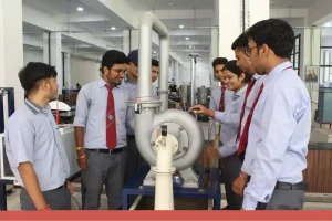 Engineering students in light blue shirts and red ties stand around a large industrial pipe/turbine apparatus in a lab, inspecting and discussing its components.