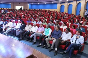 Audience seated in a large conference hall with red chairs, many people attentively facing a stage and projector screen at the front.
