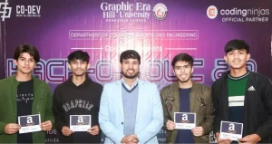 Five young men posing on stage at a hackathon event, holding Amazon cards, with a purple university banner in the background.