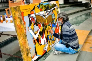 Man with gray hair and glasses painting a colorful abstract mural on a large panel outdoors, kneeling on the ground with painting supplies nearby.