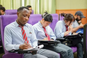 Student in a blue shirt and red tie taking notes in a lecture hall with purple seats, focused on work.