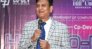 Man in a light gray checked suit and blue tie speaks into a handheld microphone on stage at a tech event backdrop with logos in purple.