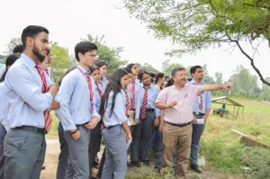 Teacher explains something to a group of students outdoors, pointing toward the field as they listen attentively.