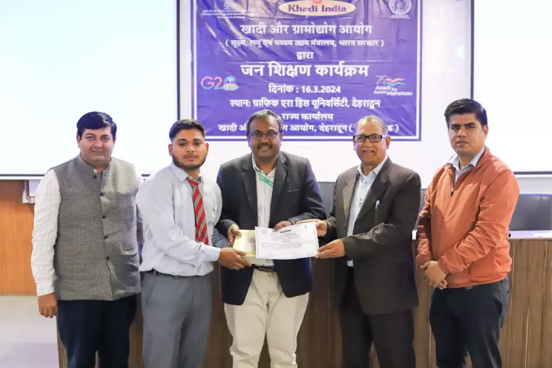 Group of men in formal attire exchanging a certificate at a stage with a blue banner in Hindi in the background.