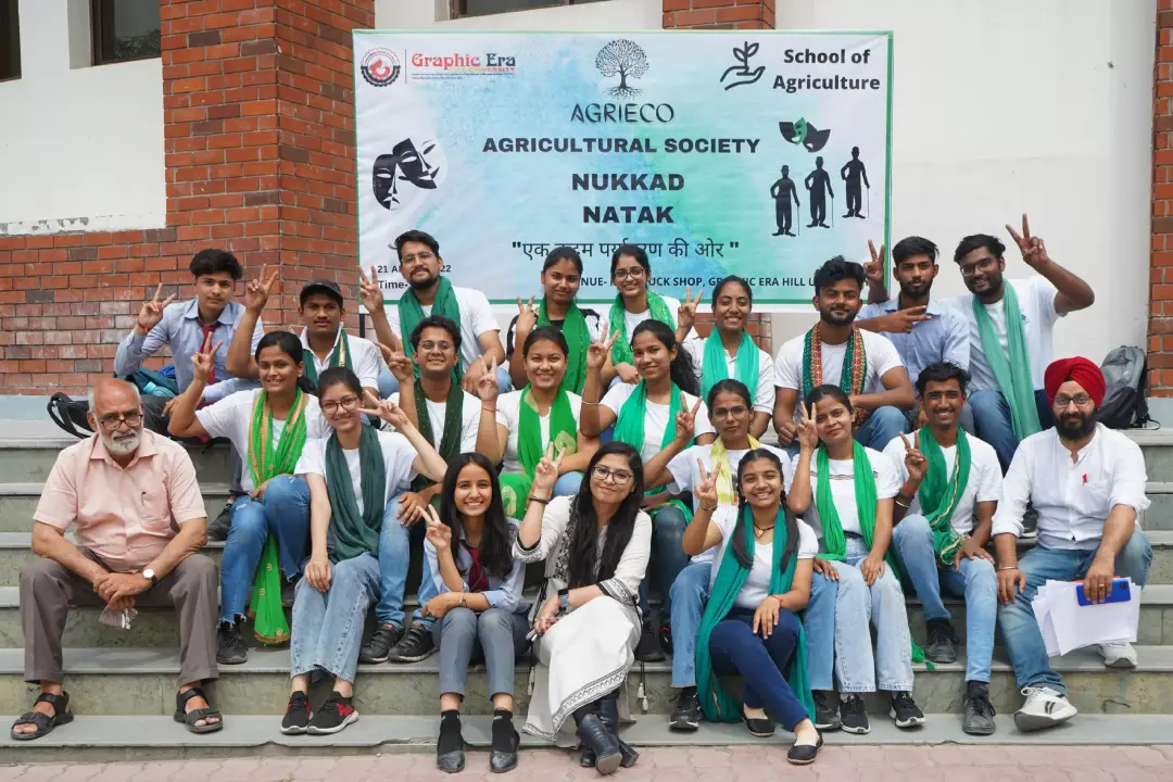 Group of students wearing green scarves posing on steps in front of a banner for Agricultural Society Nukkad Natak event, smiling and making peace signs.