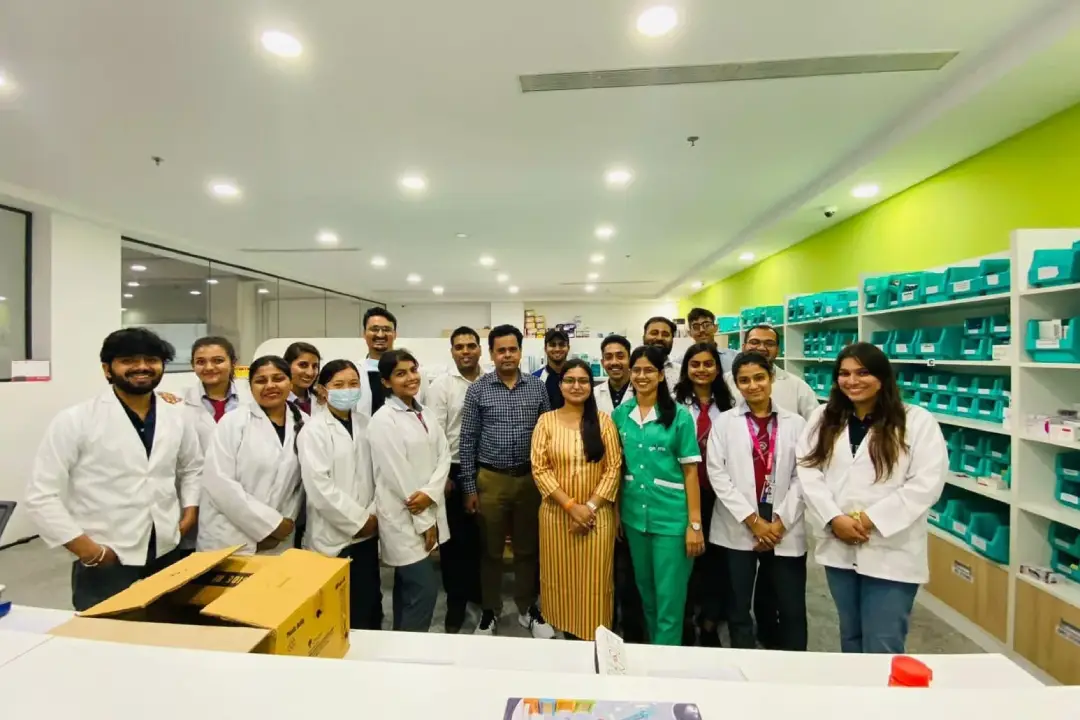 Group of healthcare workers in white coats and green scrubs posing for a team photo in a bright lab with teal bins on shelves behind them.