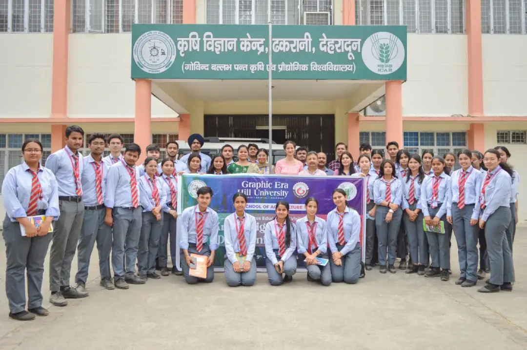 Group of students in light blue shirts and red striped ties pose outside a campus building, holding a banner for Graphic Era Hill University.