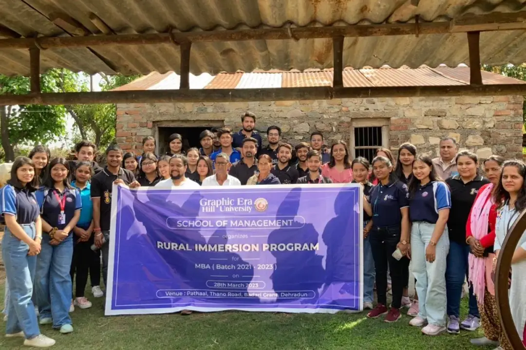 Group of students and faculty posing with a purple banner for Graphic Era Hill University's Rural Immersion Program (MBA batch 2021–2023) outdoors in front of a stone building with greenery behind.