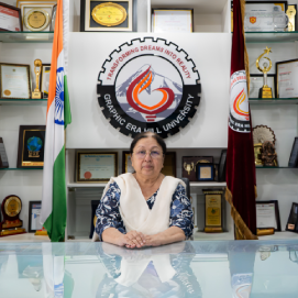 Woman seated at a glass desk in an office, with an organization banner and flags behind, surrounded by awards and plaques.