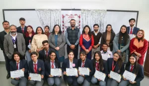 Group of students in uniforms posing for a certificate ceremony: front row kneeling with diplomas, back row of peers and teachers, banner and silver streamers behind them.