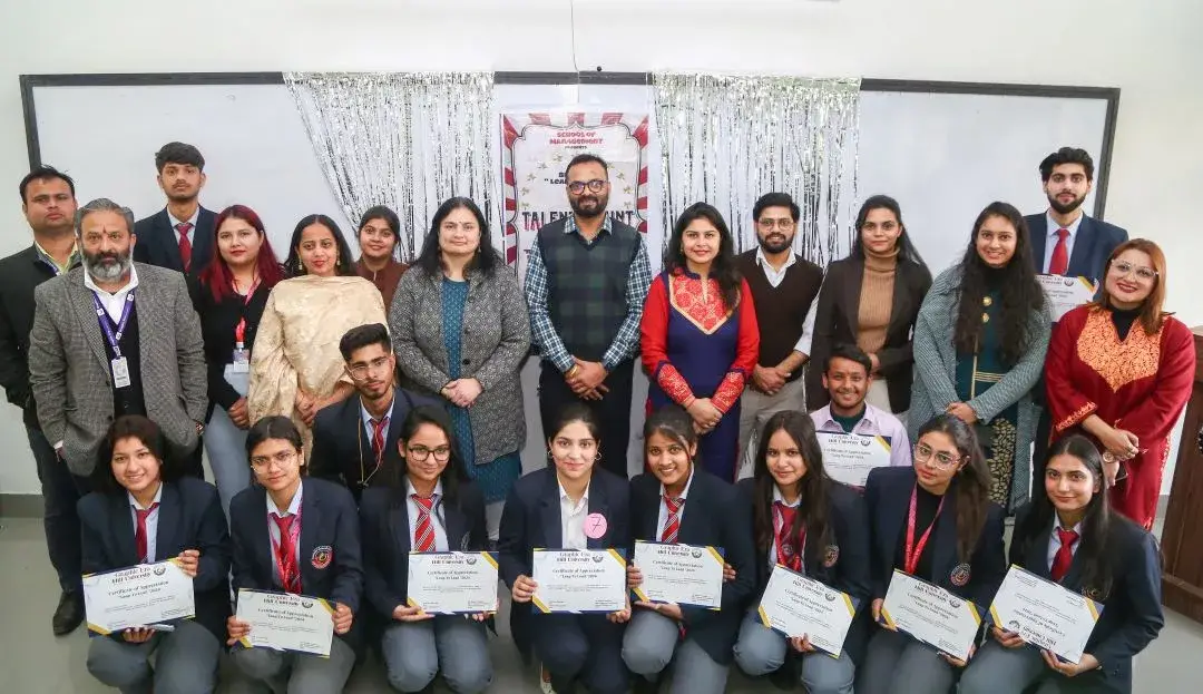 Group of students in uniforms posing for a certificate ceremony: front row kneeling with diplomas, back row of peers and teachers, banner and silver streamers behind them.