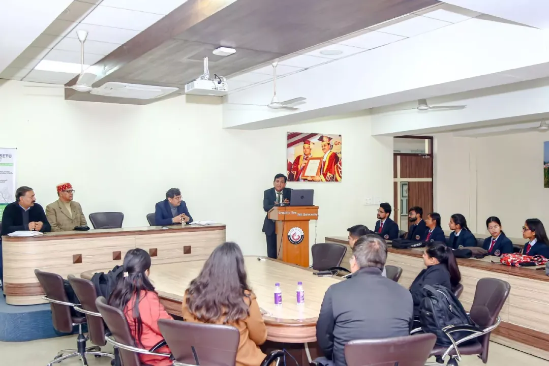 Speaker at a wooden podium delivering a presentation to a panel on the left and students in uniform on the right in a conference room.