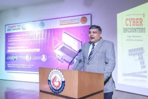 Man in a gray suit speaking at a wooden podium with a university emblem, giving a presentation at a conference with a purple event banner behind him.