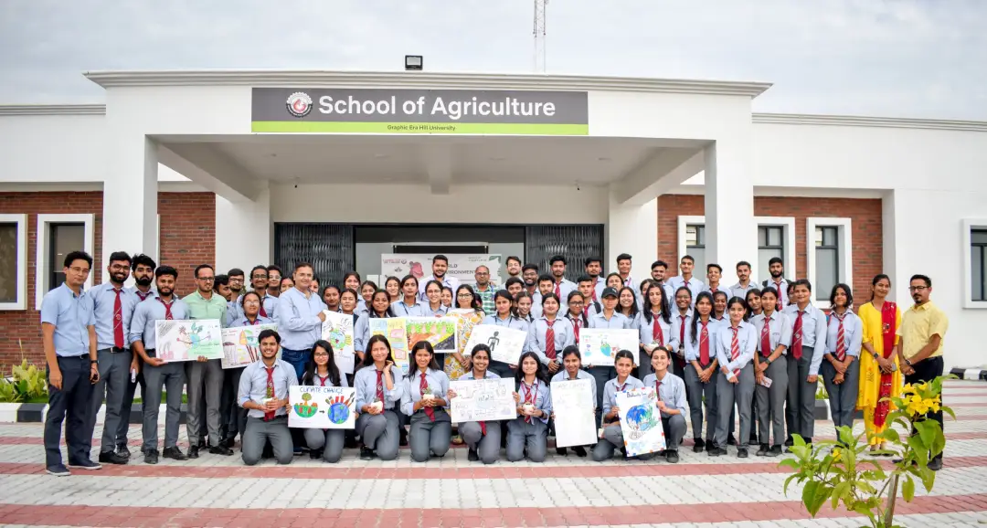Large group of students in uniform posing for a photo in front of a 'School of Agriculture' building, holding colorful posters about environmental topics.