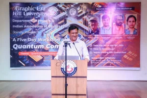 Man in a white shirt speaks from a wooden podium with a microphone, at a Quantum Computing workshop backdrop with a colorful banner.