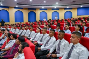 Large lecture hall filled with students in white shirts and striped ties seated in red chairs, facing the stage.