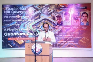 Speaker in a light shirt stands at a wooden podium with a circular logo, presenting at a Quantum Computing workshop banner in the background.