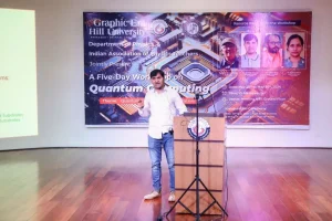 Man in a white shirt stands at a wooden podium on a stage, delivering a talk with a colorful Quantum Computing banner behind him.