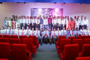 Group of students in uniforms and faculty posing on a stage in an auditorium; red theater seats visible in the foreground.