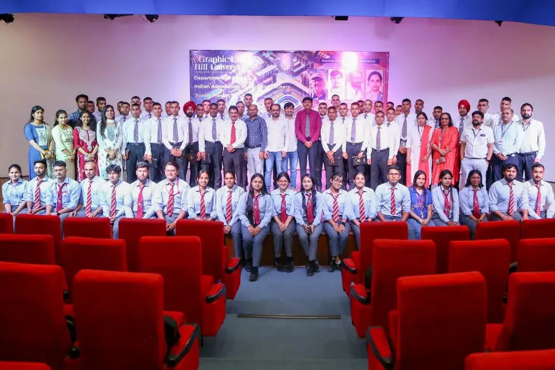 Group of students in uniforms and faculty posing on a stage in an auditorium; red theater seats visible in the foreground.